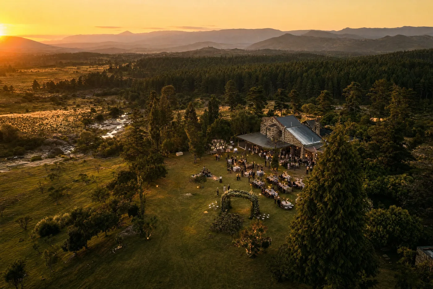 Ilustración generada con IA: vista aérea al atardecer de un evento al aire libre en una casona de piedra, con un arco de flores y mesas con luces cálidas; los invitados se ven diminutos.