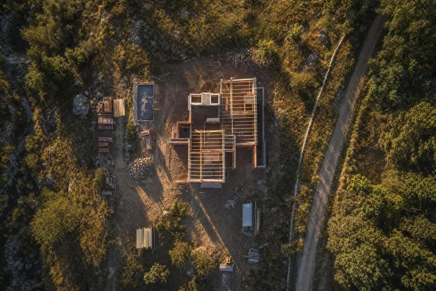 Ilustración generada con IA: vista cenital de una obra en construcción con la estructura de madera del techo, rodeada de monte serrano y junto a un camino de tierra.