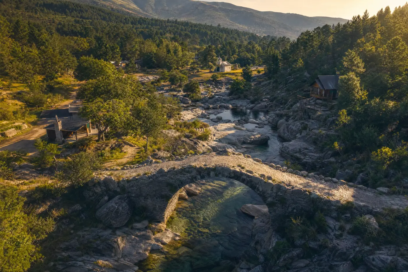 Ilustración generada con IA: vista aérea de un paraje serrano con un puente de piedra sobre un arroyo de aguas claras, algunas cabañas y una capilla entre pinares.
