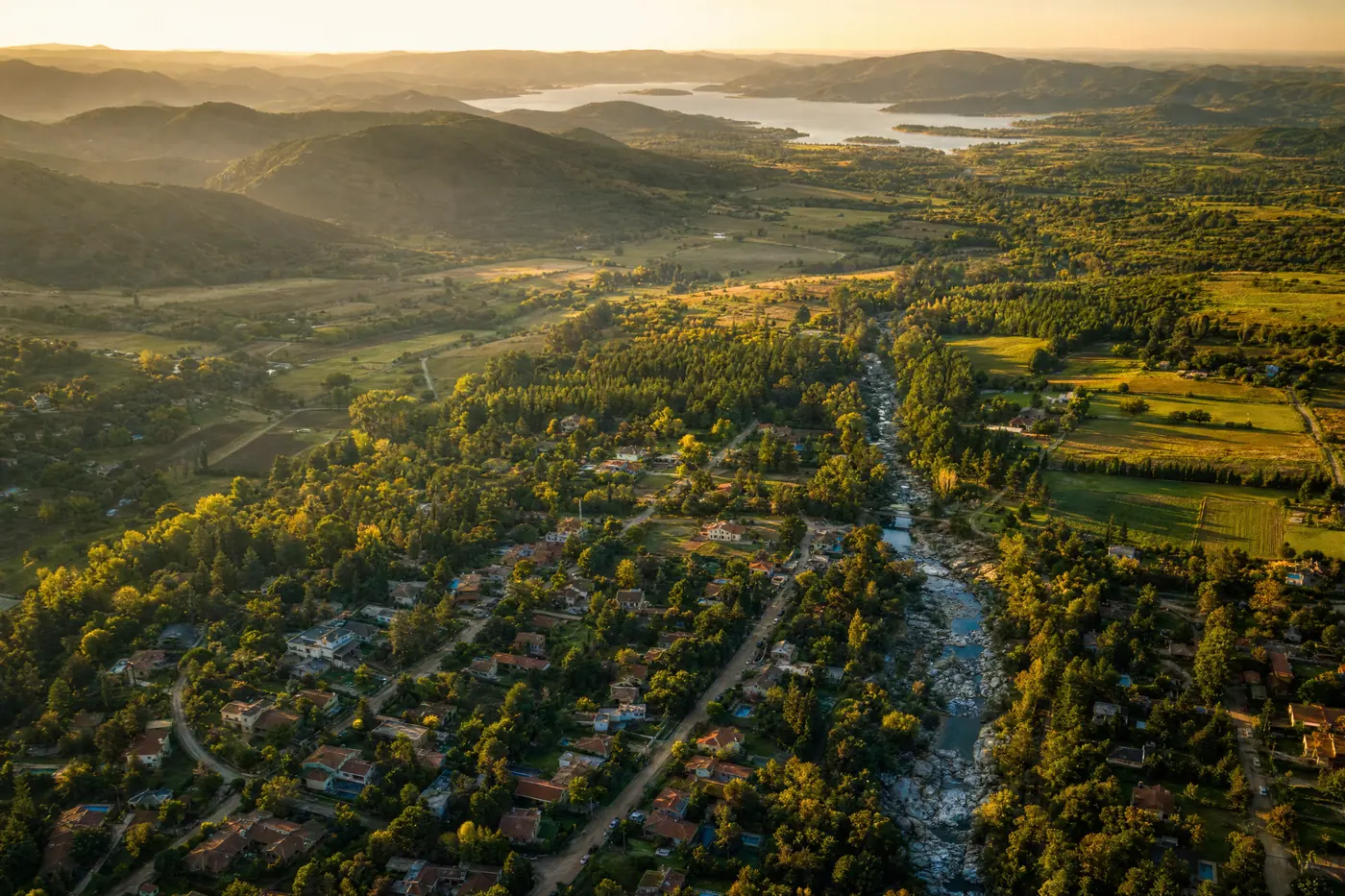 Ilustración generada con IA: vista aérea de un valle serrano con un pueblo de techos de teja junto a un arroyo, campos, pinares y un lago a lo lejos, a la hora dorada.