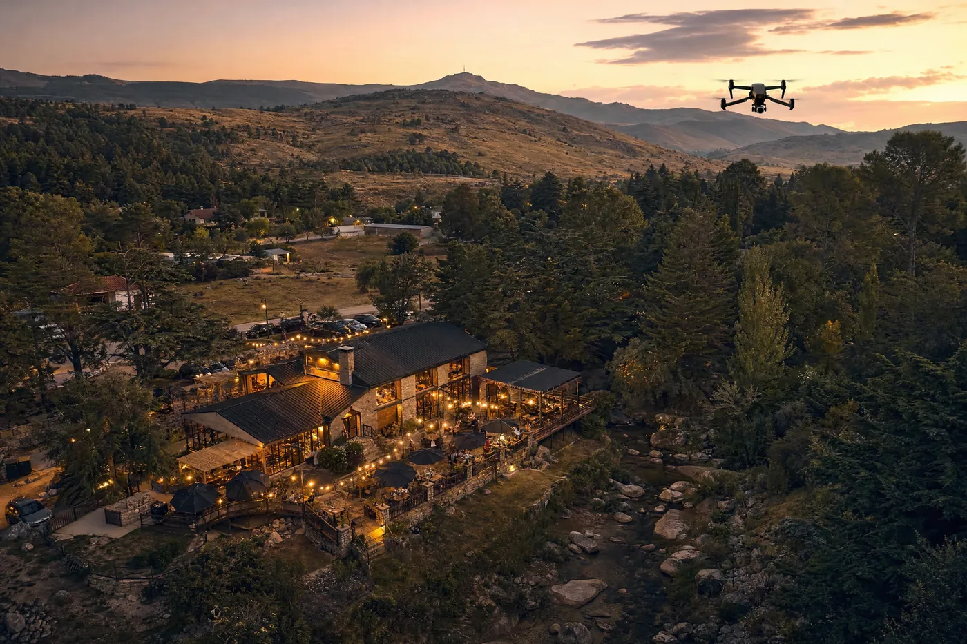 Ilustración generada con IA: vista aérea al anochecer de un restaurante de pueblo serrano con terraza iluminada con guirnaldas, rodeado de árboles, y un drone en el cielo.