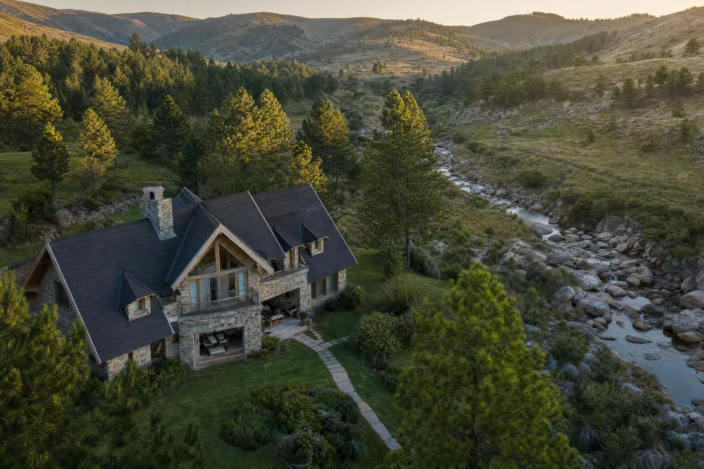 Ilustración generada con IA: vista aérea de una casa de estilo alpino de piedra y madera, con techo a dos aguas y jardín entre pinos, junto a un arroyo de piedras en las sierras.